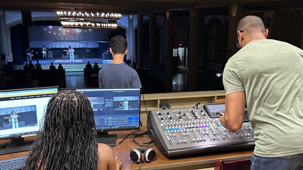 A view from behind a control room showing two individuals operating sound equipment while a person is speaking on stage with an audience in the background.